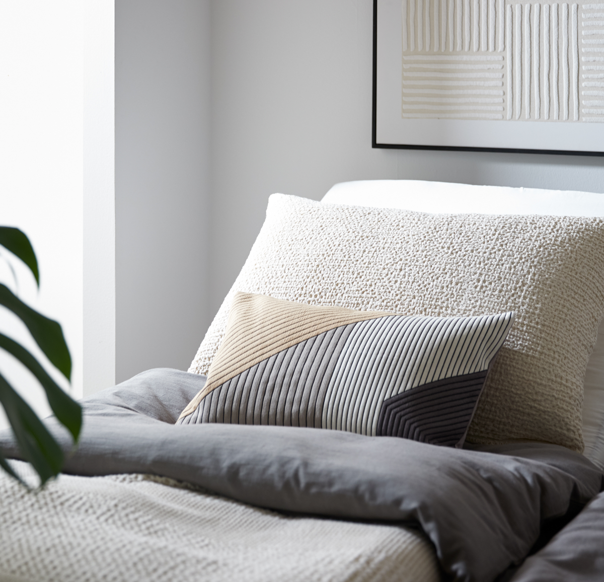 Nærbillede af en redt seng med stor tekstureret pude og aflang pude med ribbet, geometrisk design i beige, grå og hvid i et lyst, minimalistisk soveværelse.