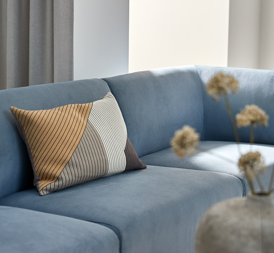 Blå hjørnesofa med blød polstring og en stribet pyntepude i beige, grå og hvid; blødt dagslys og en sløret vase med tørrede blomster i forgrunden.