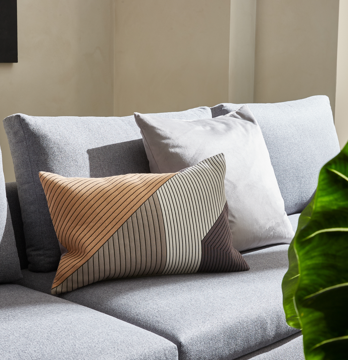 Lysegrå sofa med to puder – en lysgrå og en rektangulær med geometrisk mønster i beige, grå og brun – i solbelyst stue med grøn plante i forgrunden.
