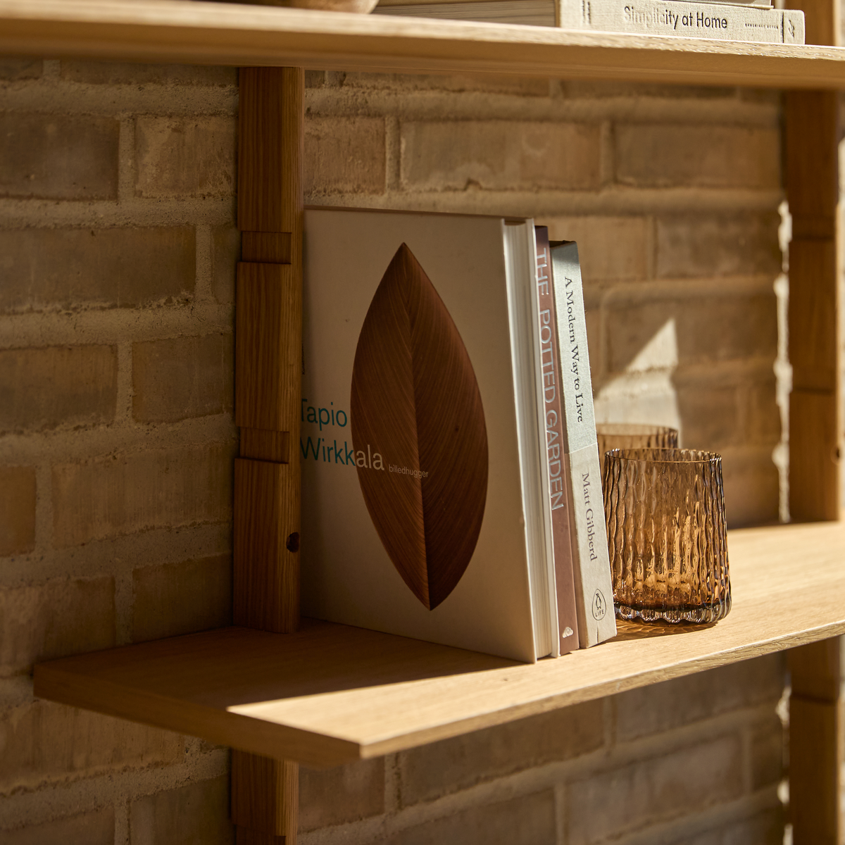 Træhylde mod murstensvæg med bøgerne ”Tapio Wirkkala”, ”The Potted Garden”, ”A Modern Way to Live” af Matt Gibberd og ”Simplicity at Home”, ved siden af et ravfarvet glas i varmt dagslys.