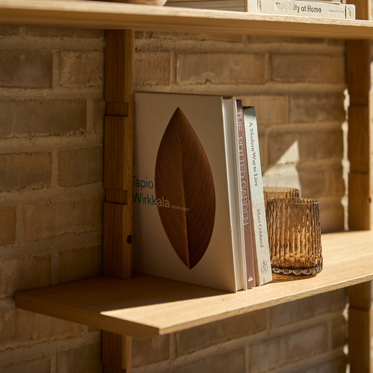 Træhylde mod murstensvæg med bøgerne ”Tapio Wirkkala”, ”The Potted Garden”, ”A Modern Way to Live” af Matt Gibberd og ”Simplicity at Home”, ved siden af et ravfarvet glas i varmt dagslys.