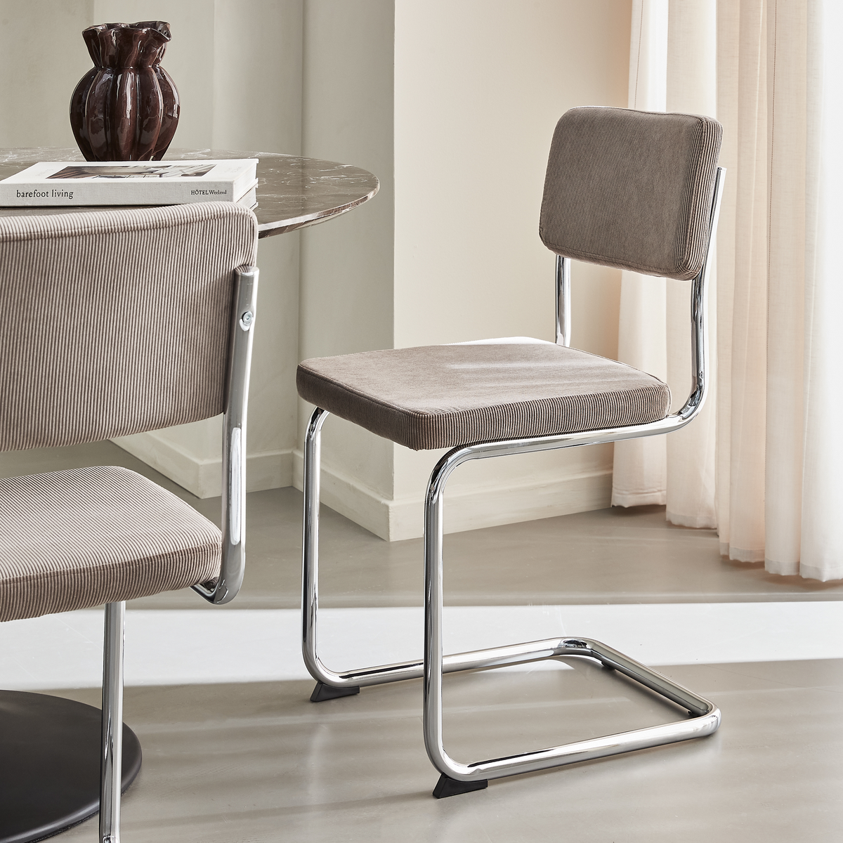 Moderne cantilever-spisestol i beige fløjl med forkromet stel ved siden af et rundt marmorbord med en mørk vase og bøgerne “barefoot living” og “HOTEL Weekend”.