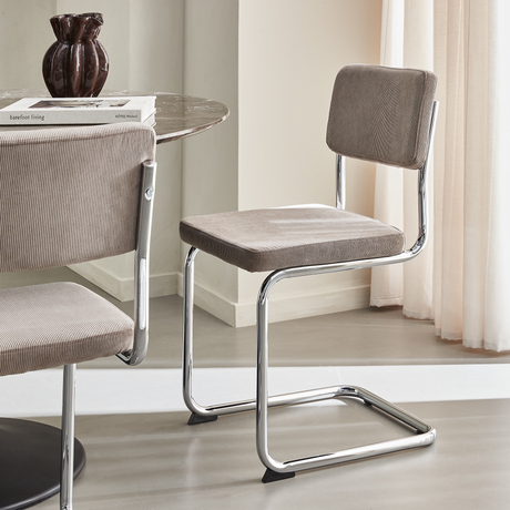 Moderne cantilever-spisestol i beige fløjl med forkromet stel ved siden af et rundt marmorbord med en mørk vase og bøgerne “barefoot living” og “HOTEL Weekend”.