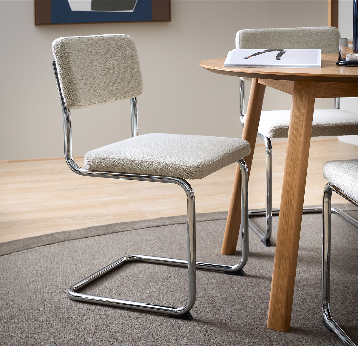 Beigefarvet cantilever-stol med kromstel ved et rundt egetræsbord på et gråt tæppe i en minimalistisk spisestue.