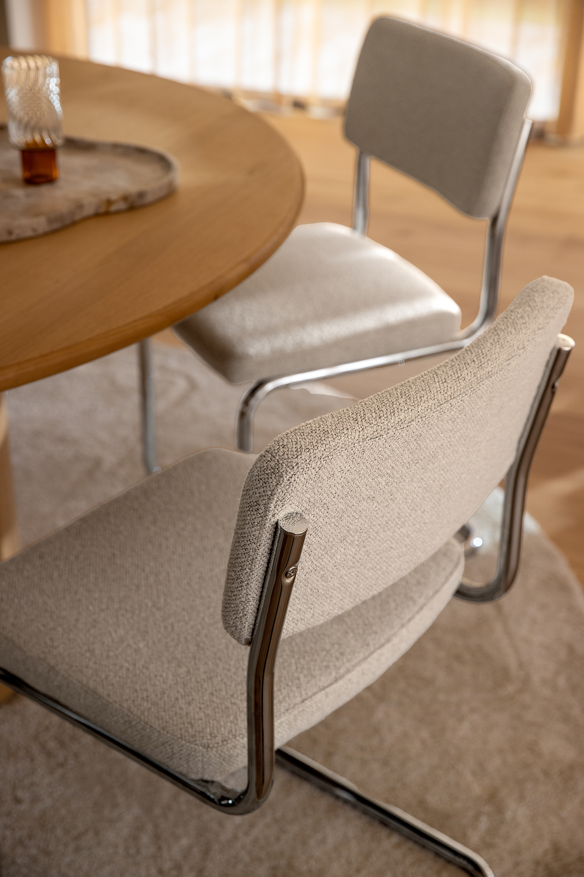 Nærbillede af to beige polstrede cantilever-stole med kromstel ved et rundt spisebord i træ, i blødt dagslys på et neutralt tæppe.