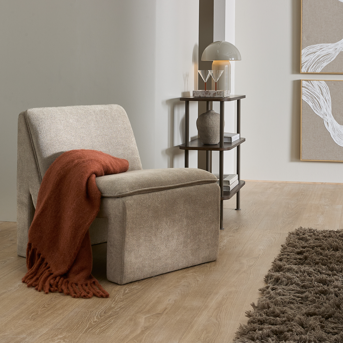 Beige armløs lænestol med rustfarvet plaid ved siden af et mørkt sidebord med kuppellampe, vase og bøger på lyst trægulv med brunt rygtæppe i en lys stue.