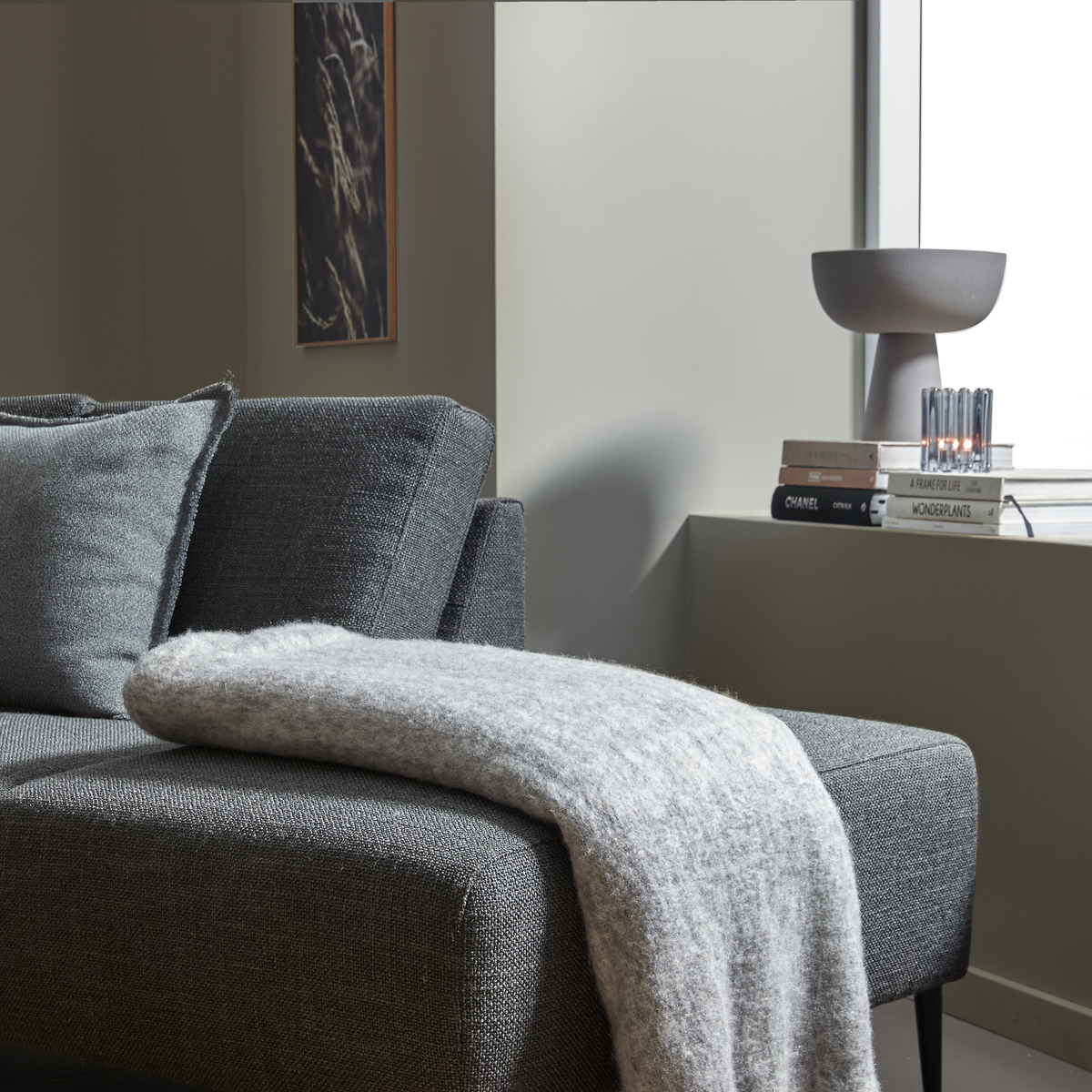 Nærbillede af en grå sofa med lysgråt tæppe; på vindueskarmen står en grå bordlampe, et lille lys og en bogstak med titlerne CHANEL, A FRAME FOR LIFE og WONDERPLANTS.
