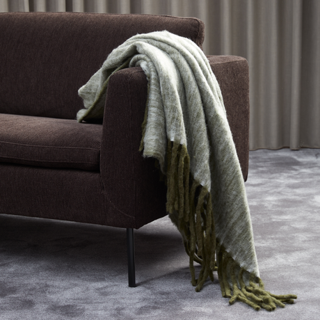 Grøn‑grå plaid med frynser hænger over armlænet på en mørkebrun sofa, med gråt tæppe og gardiner i baggrunden.