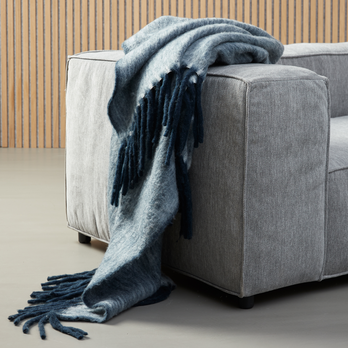 Blå meleret plaid med frynser draperet over armlænet på en grå sofa.