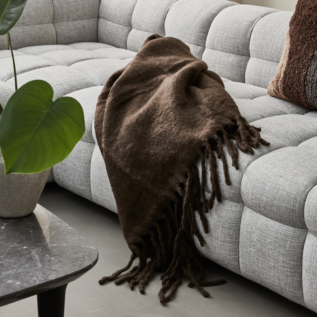 Brunt uldtæppe med frynser er lagt over en lysegrå, quiltet sofa; grøn potteplante og mørk marmorbordplade i forgrunden.