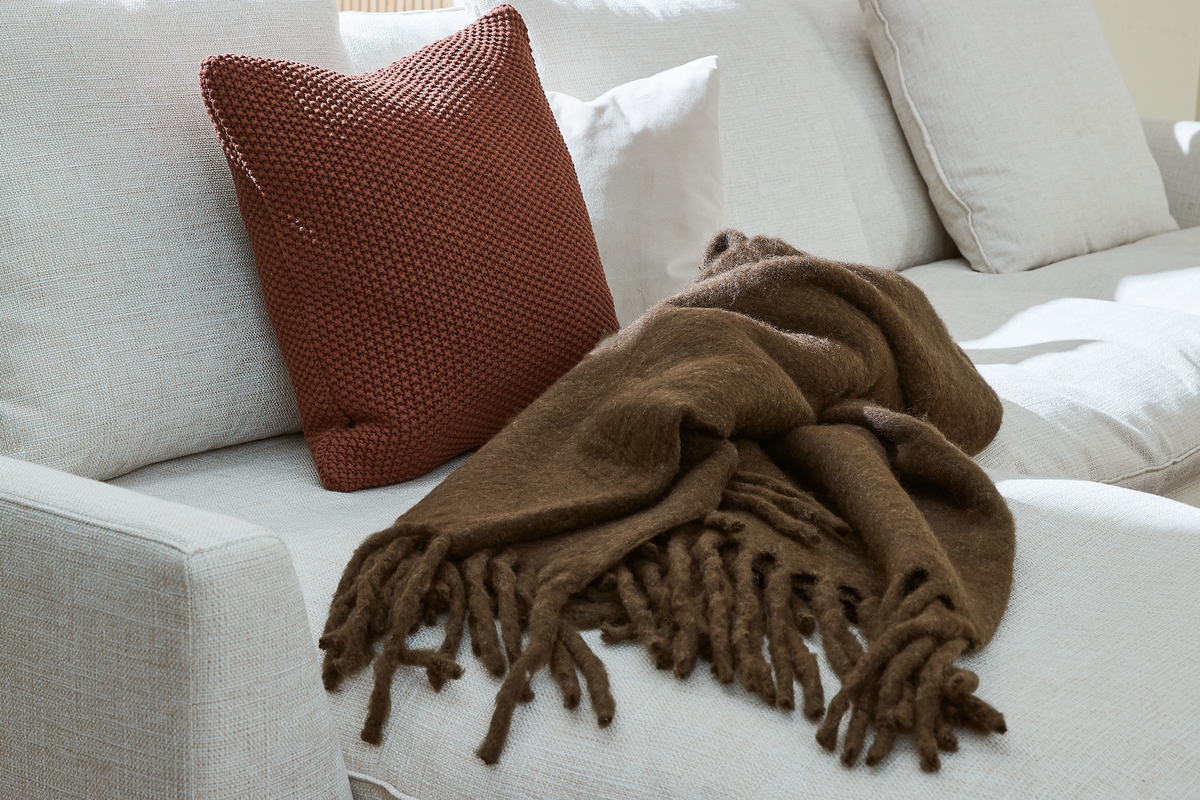 Lys sofa med rustfarvet strikket pude og brun plaid med frynser lagt over sædet i dagslys.