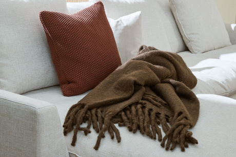 Lys sofa med rustfarvet strikket pude og brun plaid med frynser lagt over sædet i dagslys.