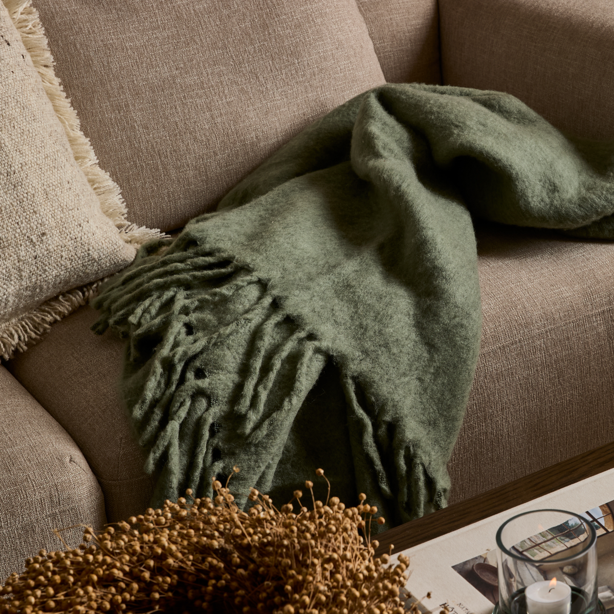 Nærbillede af et blødt grønt plaid med frynser lagt over en beige sofa med en tekstureret pude; på sofabordet står et stearinlys i glas og et magasin.
