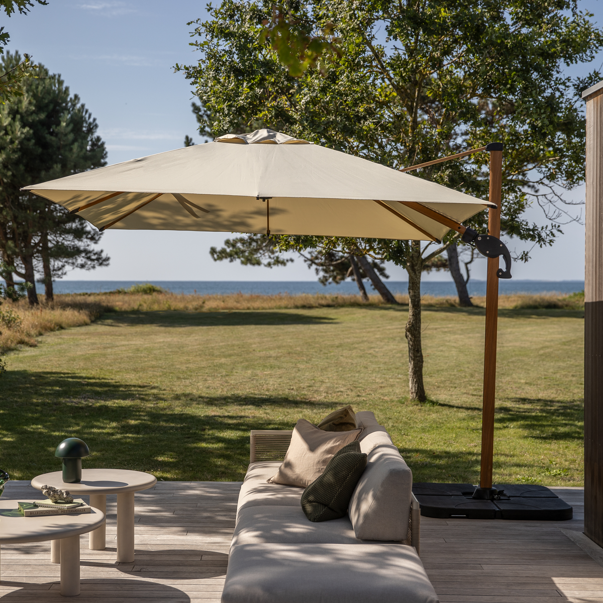 Udendørs lounge på træterrasse med beige sofa, runde sideborde og stor fritstående parasol med udsigt over græsplæne, træer og hav i sollys.