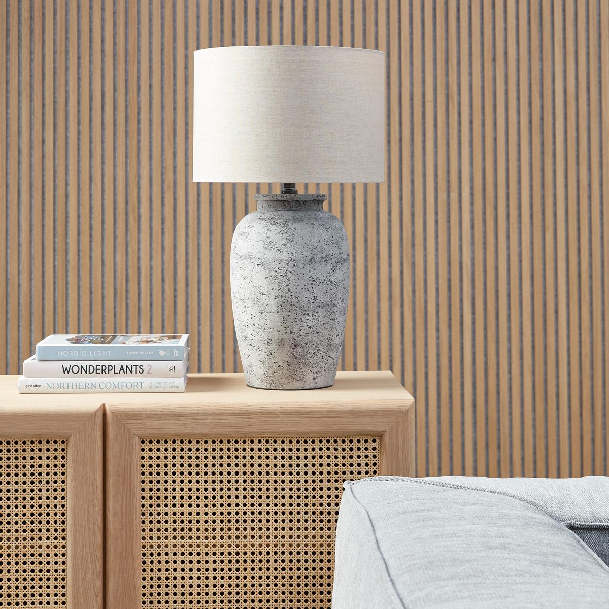 Bordlampe i beton med lys stofskærm på et skab med rattanfronter, ved siden af en stak bøger med titlerne Nordic Light, Wonderplants 2 og Northern Comfort.