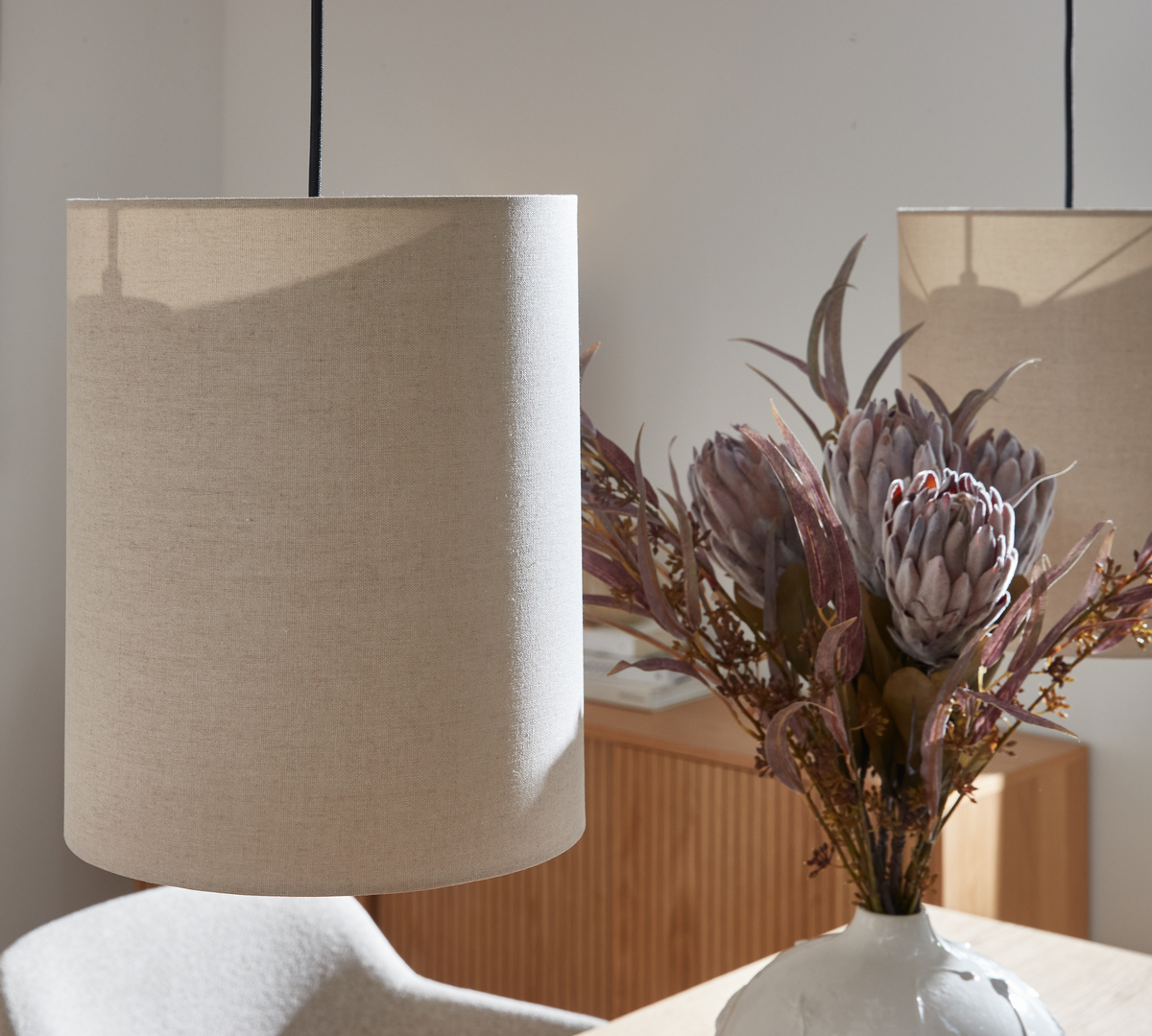 Nærbillede af en beige cylinderformet pendellampe over et bord, med en hvid vase med protea-blomster i et lyst, neutralt hjem.