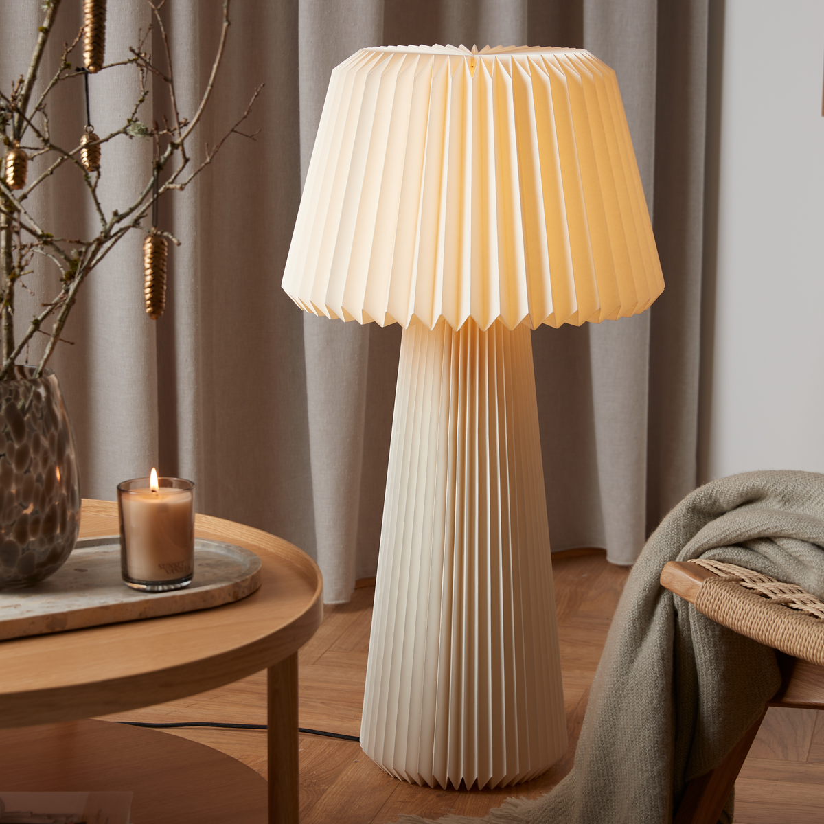 Plisseret gulvlampe med varmt hvidt lys ved siden af rundt sofabord i træ med tændt lys og vase med grene, grå gardiner i baggrunden og et plaid på stolen.