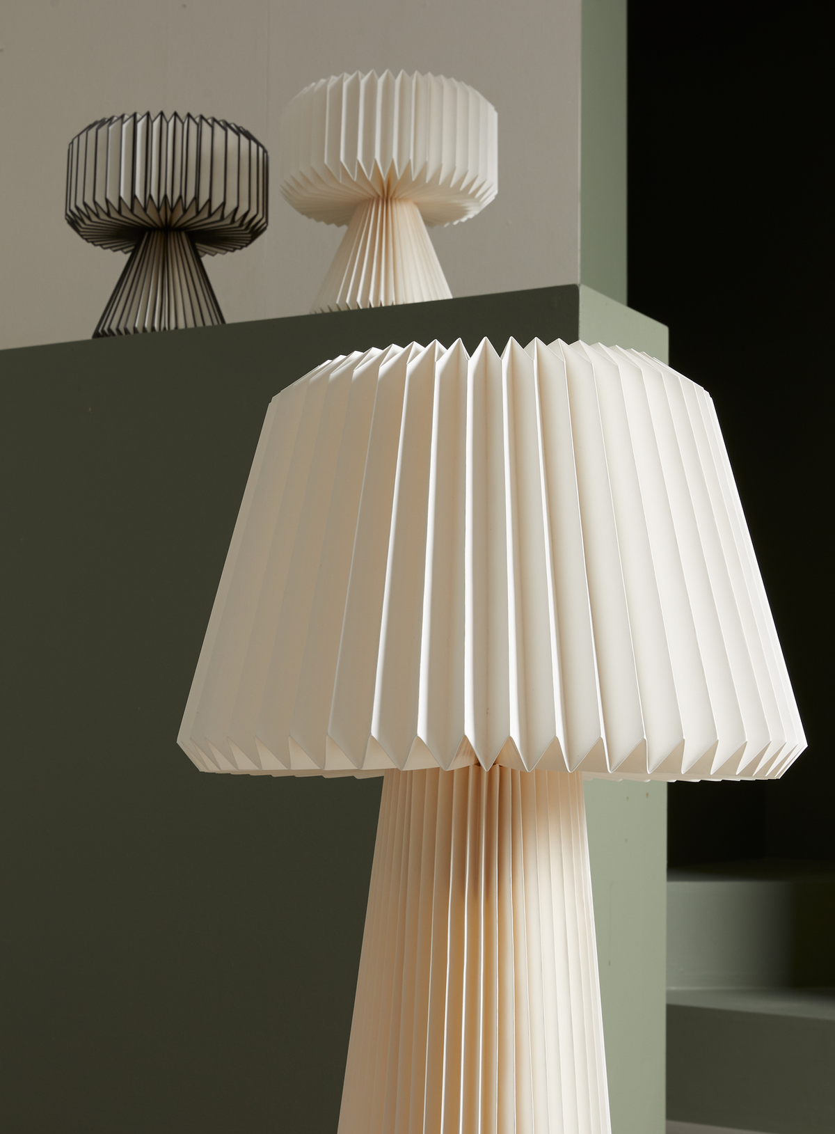 Plisserede bordlamper i beige og hvid mod grøn baggrund; en stor i forgrunden og to mindre i baggrunden.