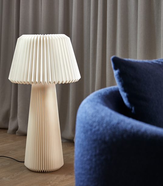Beige plisseret bordlampe ved grå gardiner med en blå sofa i forgrunden.