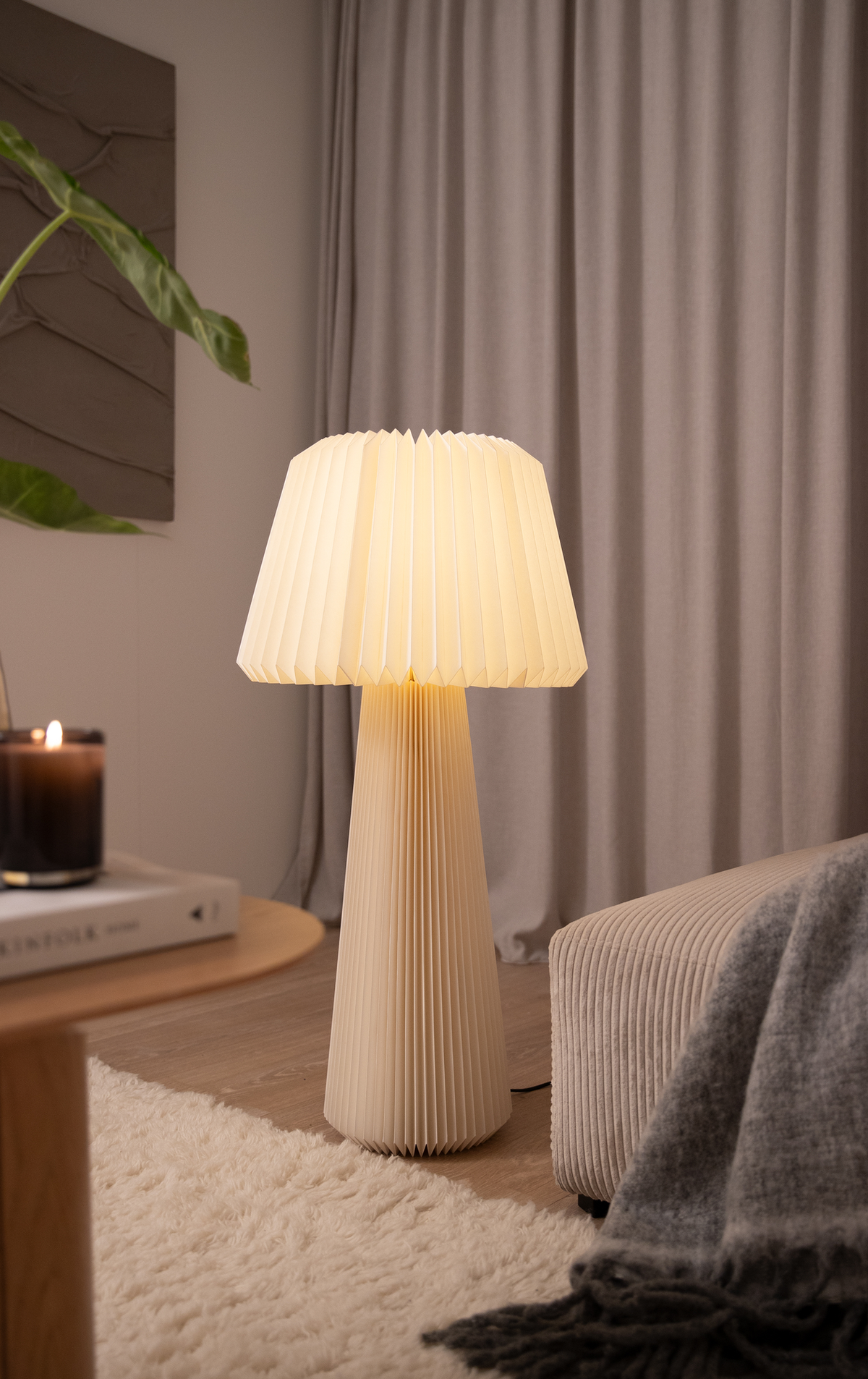 Hygelig stue med en varmglødende plisseret gulvlampe ved siden af en sofa, et tændt lys og en bog på et lille bord, gardiner i baggrunden og et blødt tæppe på gulvet.