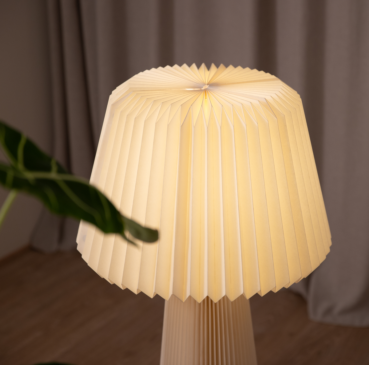 Plisseret bordlampe med varmt lys ved siden af gardin og grøn plante.