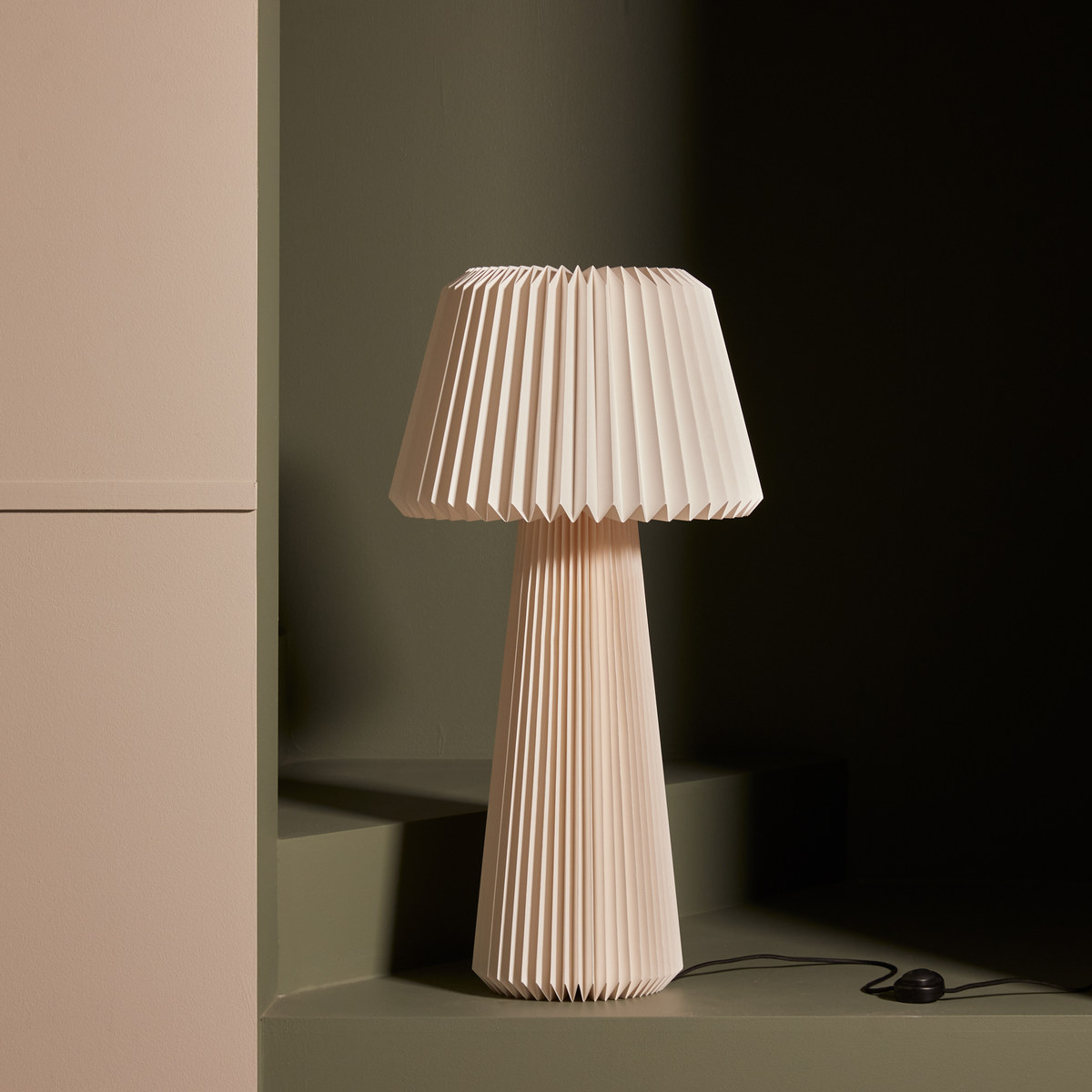 Beige bordlampe med plisseret skærm og fod i et minimalistisk studiomiljø, med synlig ledning og fodkontakt.