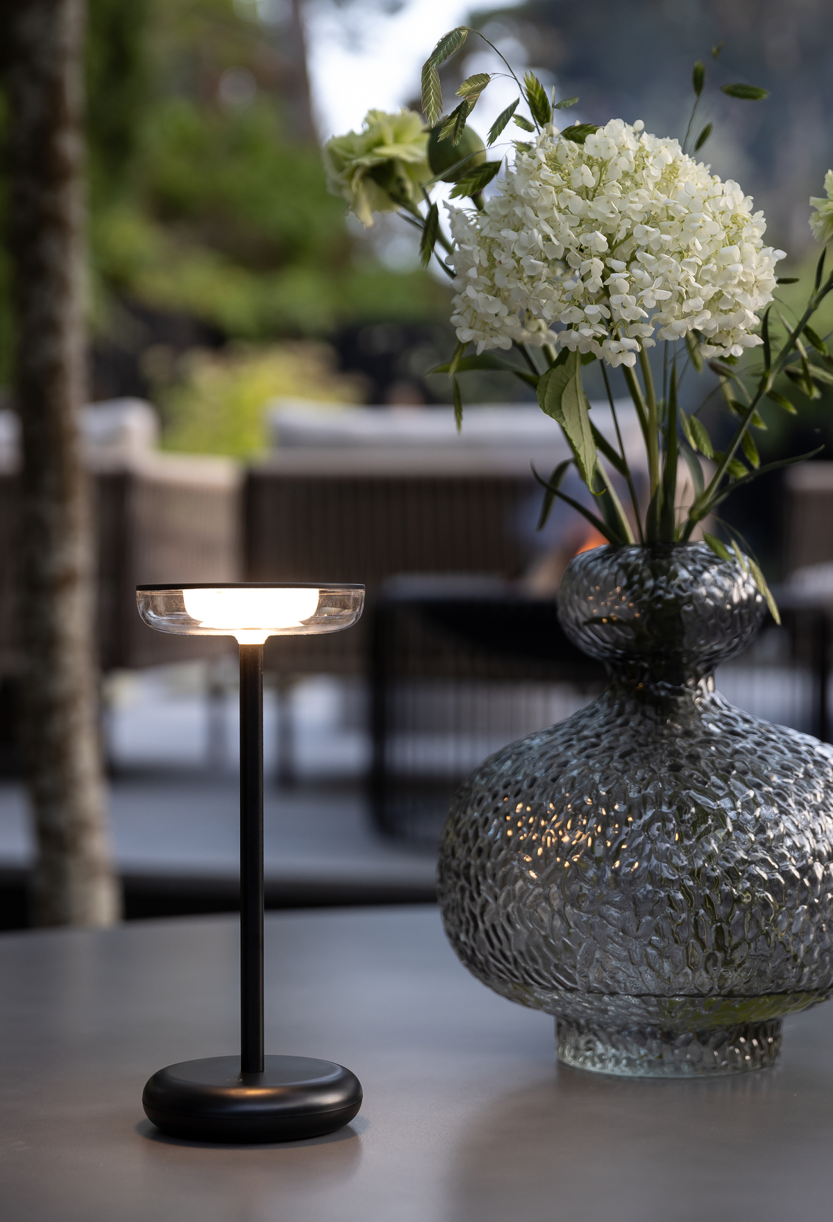Tændt, bærbar sort bordlampe ved siden af en struktureret glasvase med hvide blomster på en terrasse.