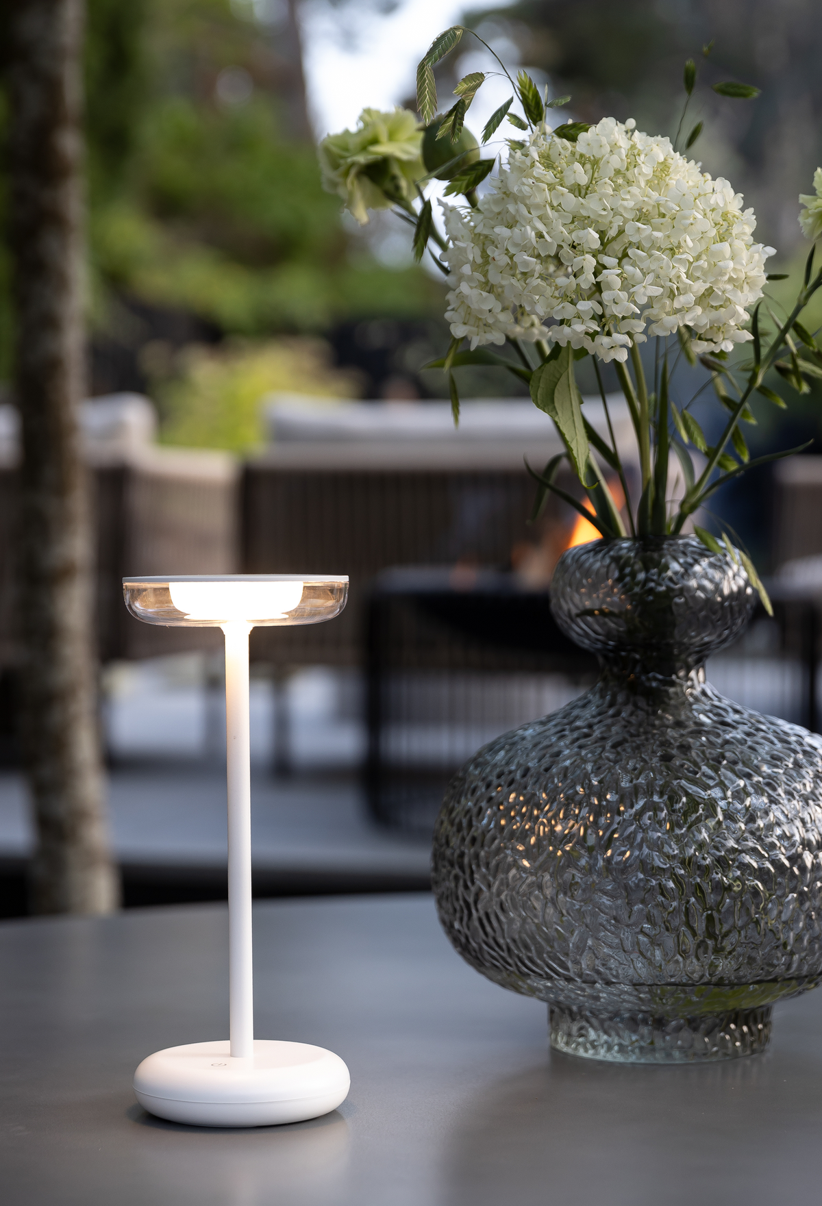 Bærbar hvid bordlampe tændt ved siden af en tekstureret glasvase med hvide blomster på et udendørs bord på en terrasse.