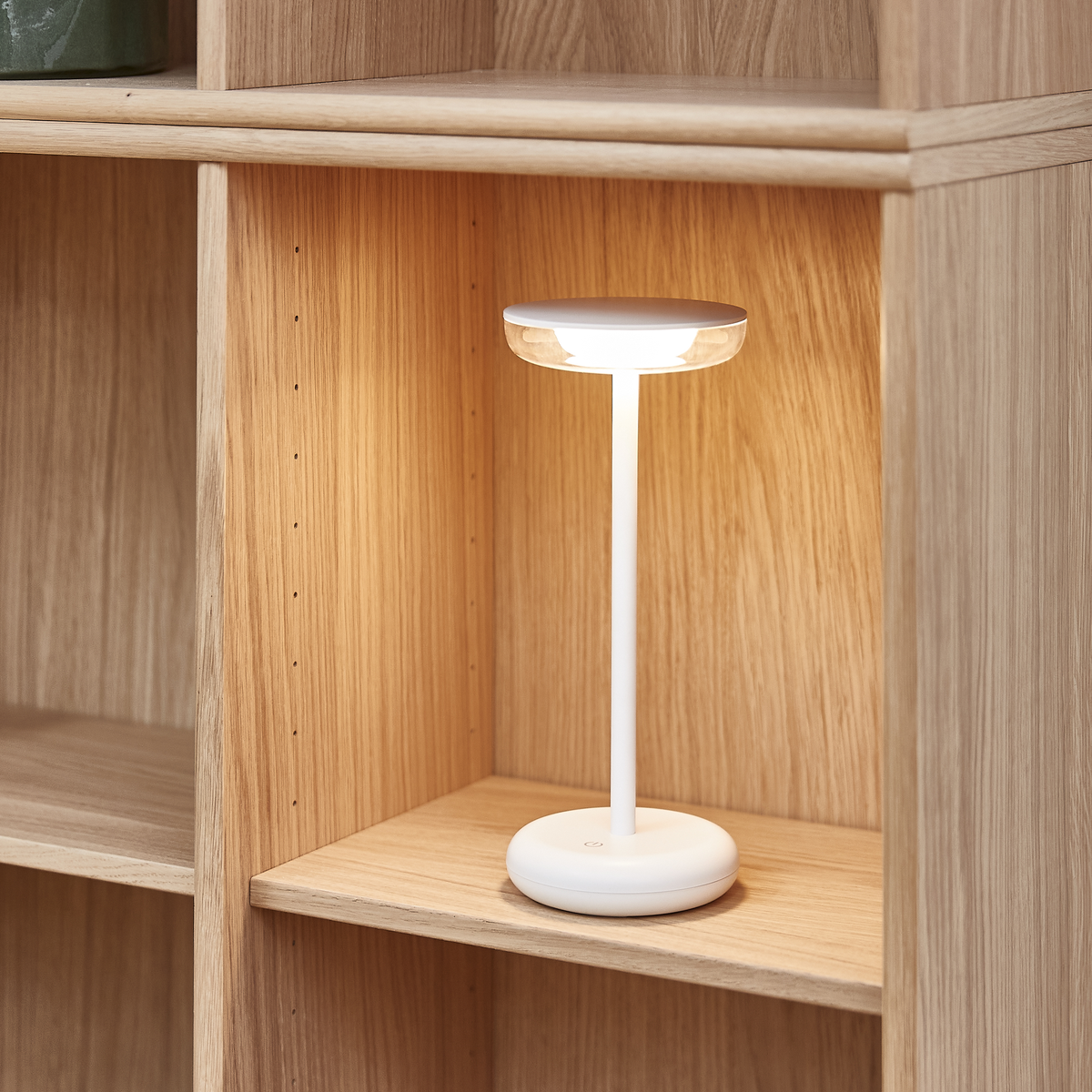 Hvid bordlampe, der lyser på en hylde i lyst egetræ.