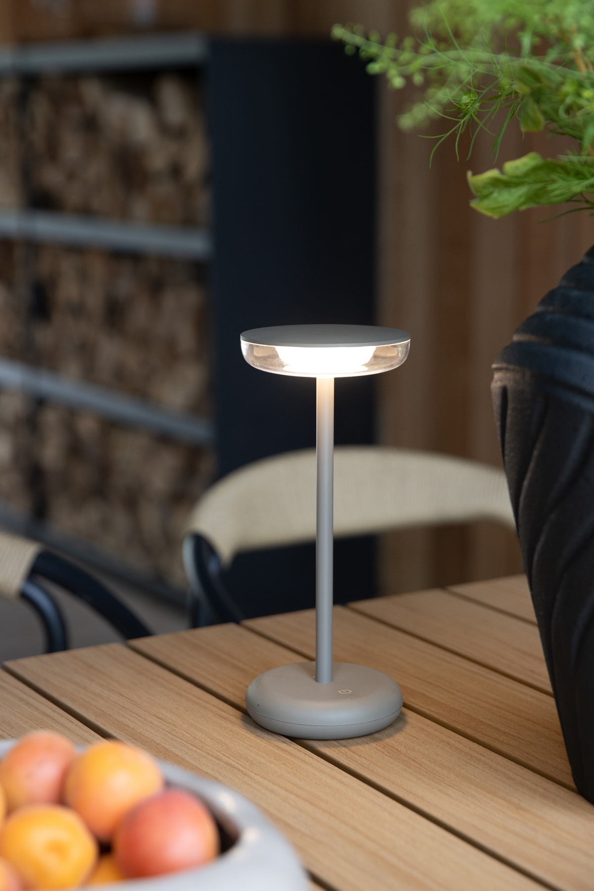 Bærbar bordlampe i mat grå med rund fod med strøm-symbol, tændt på et træbord udendørs ved siden af grøn plante.