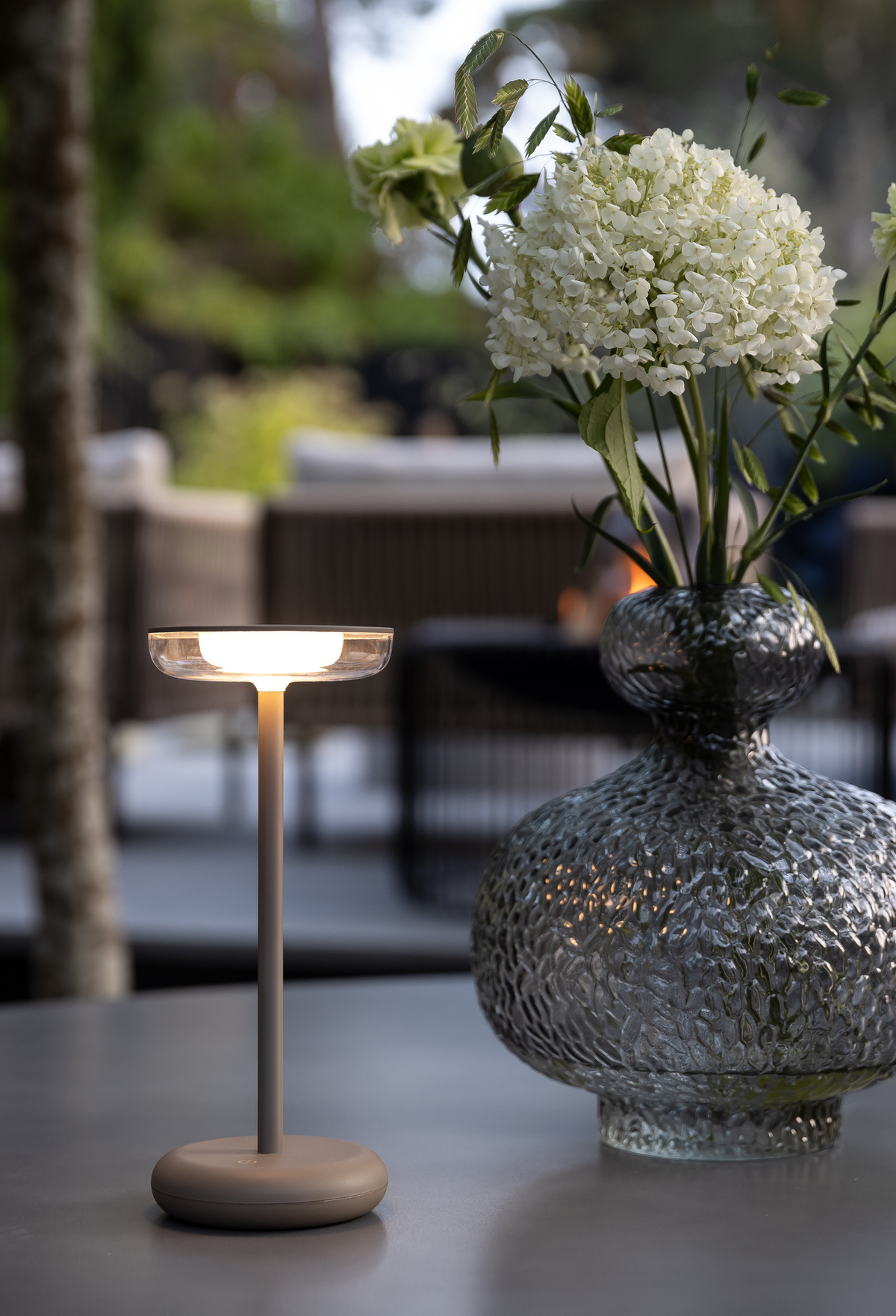 Bærbar LED-bordlampe tændt ved siden af en struktureret glasvase med hvide blomster på et udendørs bord.