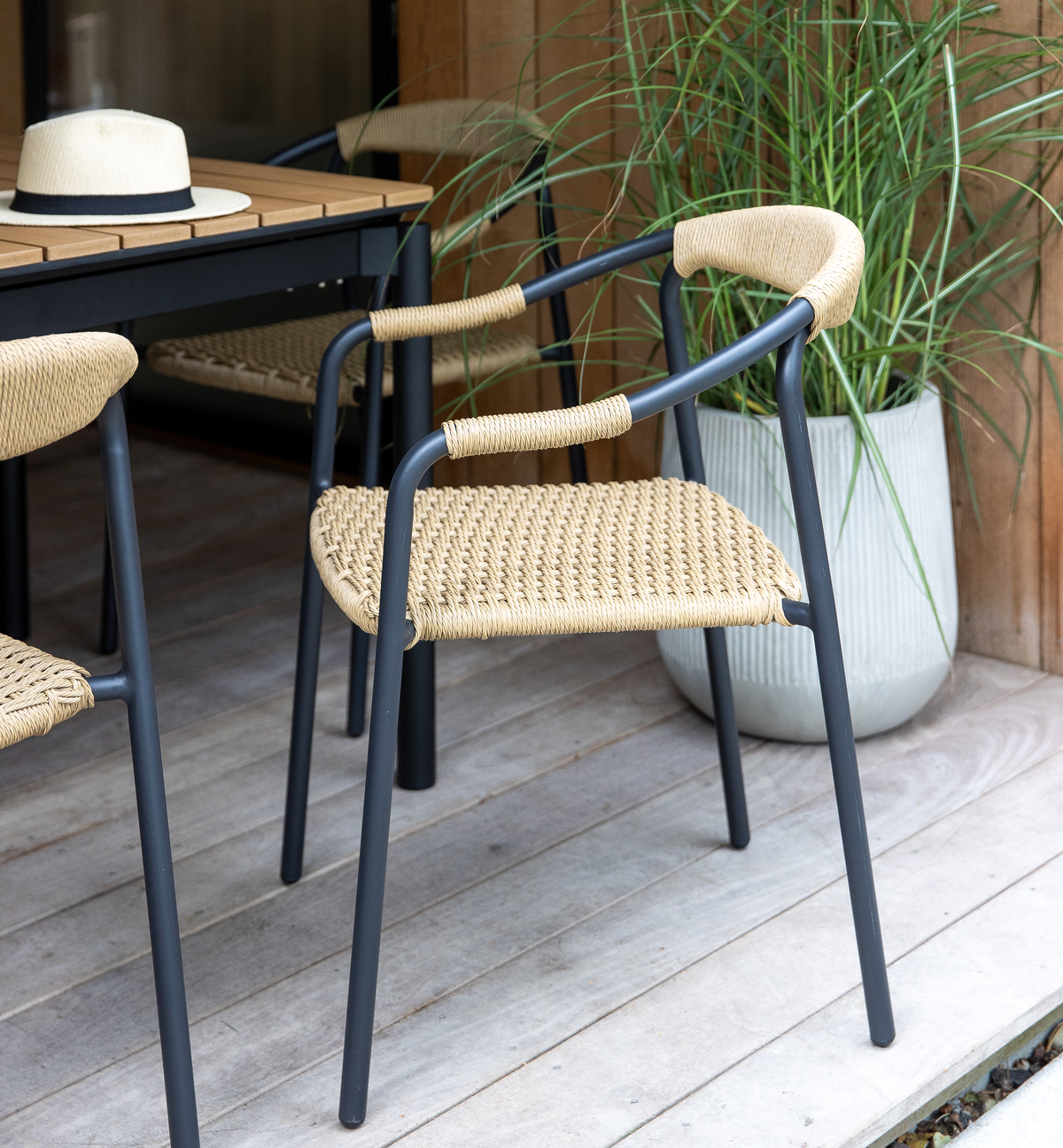 Udendørs stol med sort metalstel og naturfarvet flettet sæde og ryg ved et træbord på terrasse; grøn plante i krukke i baggrunden.