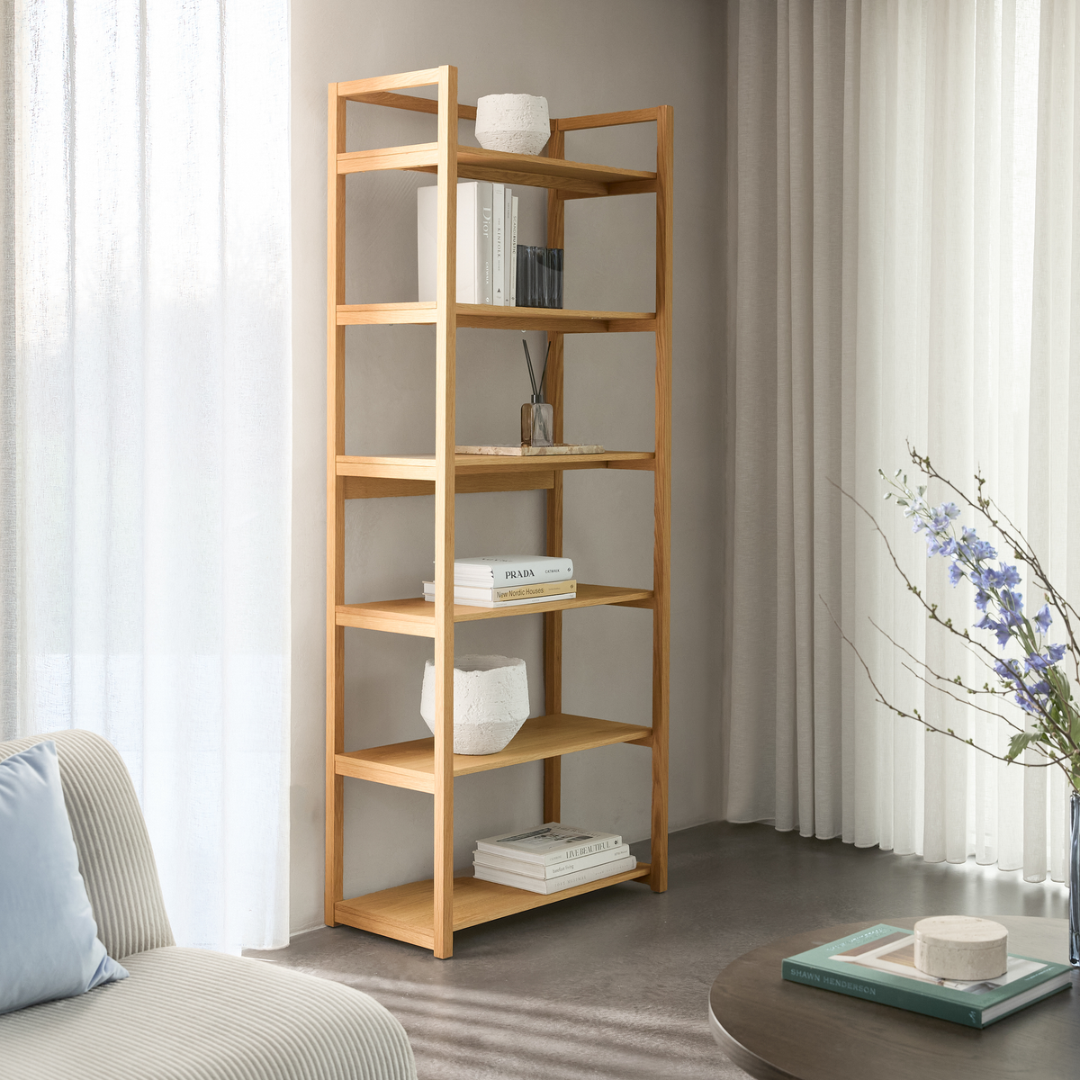 Lyst stueinteriør med åben reol i eg, pyntet med få bøger (bl.a. ”PRADA” og ”New Nordic Houses”) og hvide keramikkar; ved siden af sofa, lette gardiner og et sofabord med vase og blomster.