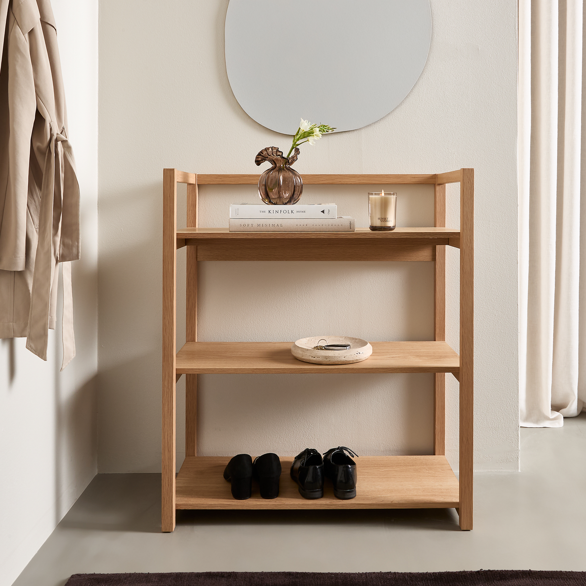 Lys entréscene med en reol i egetræ under et organisk formet spejl; øverste hylde har en glasvase med en blomst, et stearinlys og bøgerne The Kinfolk Home og Soft Minimal, nederst står sorte sko.
