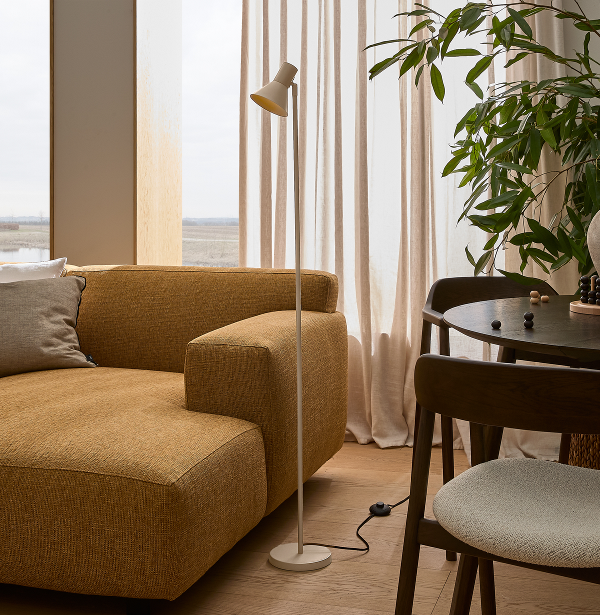 Stue med sennepsgul sofa, hvid gulvlampe og rundt træbord ved lyse gardiner og grøn plante.