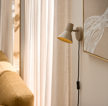 Beige væglampe med sort ledning og afbryder over en gyldenbrun sofa ved lyse gardiner i stue med blødt dagslys.