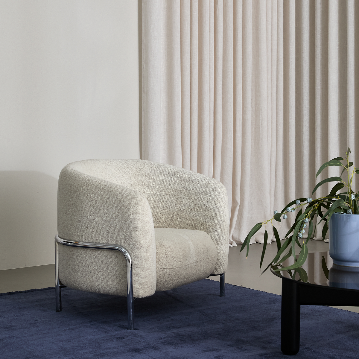 Cremefarvet lænestol i bouclé med kromben på mørkeblåt tæppe ved siden af sort sofabord med grøn plante, foran beige gardiner.