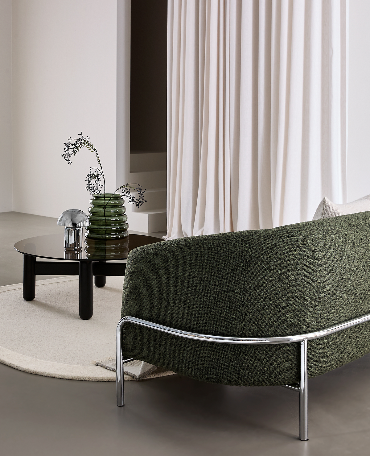 Moderne stue med mørkegrøn buet sofa med kromben, rundt sort sofabord med røgfarvet glas, grøn riflet vase med grene og lille krombordlampe på lys rund måtte foran lyse gardiner.