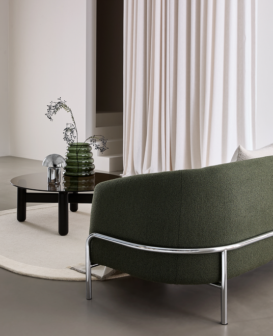 Moderne stue med mørkegrøn buet sofa med kromben, rundt sort sofabord med røgfarvet glas, grøn riflet vase med grene og lille krombordlampe på lys rund måtte foran lyse gardiner.