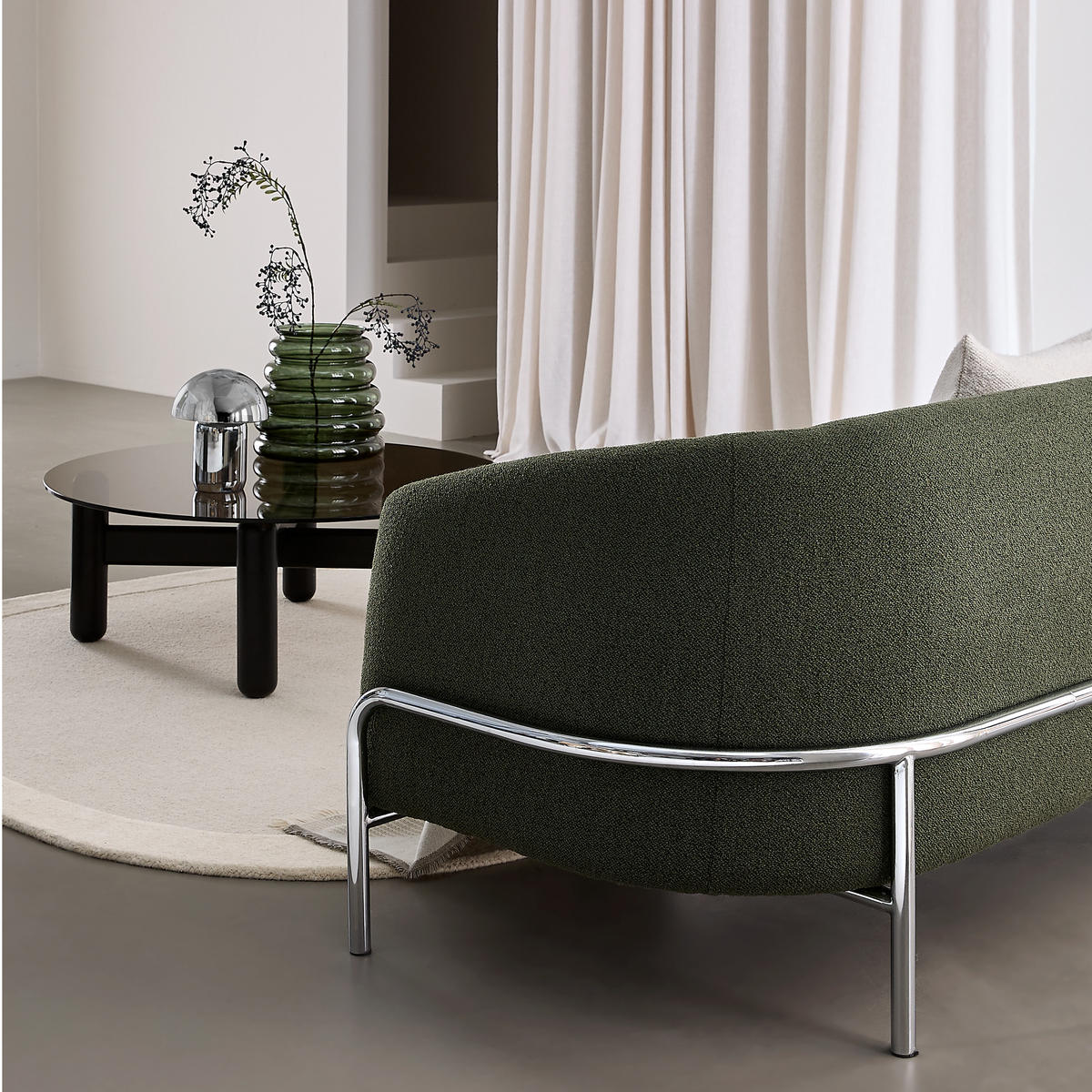 Moderne stue med mørkegrøn sofa med kromstel, lavt sort sofabord med mørk glasplade, grøn glasvase med grene og en lille krom svampelampe på et lyst rundt tæppe foran lyse gardiner.
