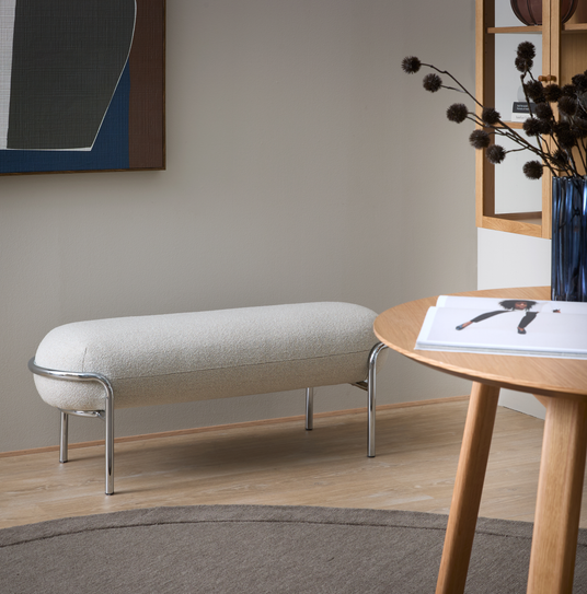 Lyst stuehjørne med beige bænk med kromben, rundt egetræsbord med åben bog og blå vase med tørrede grene samt abstrakt vægkunst.