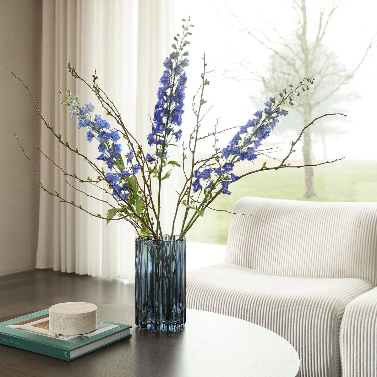 Blå glasvase med grene og blå blomster på et sofabord, ved siden af bøger og en lys lænestol ved et vindue med lette gardiner.