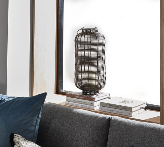 Flettet rattanlanterne med lys på vindueskarm oven på bøger – A Frame for Life, The Kinfolk Garden, Northern Comfort – ved siden af en grå sofa.