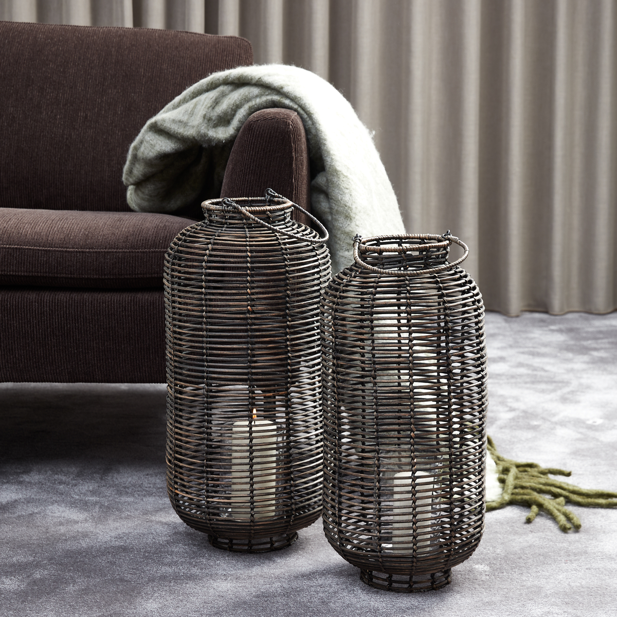To store rattan-lanterner med bloklys står på et gråt tæppe ved siden af en mørk sofa med et blødt plaid foran beige gardiner.