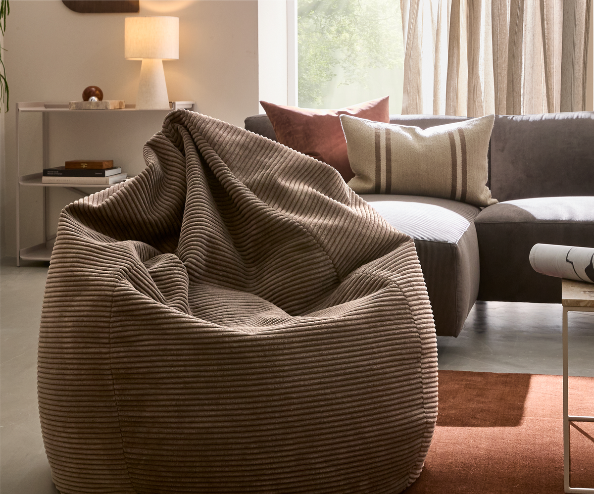 Brun fløjl-sækkestol i stue med grå sofa, puder og bordlampe i blødt dagslys.