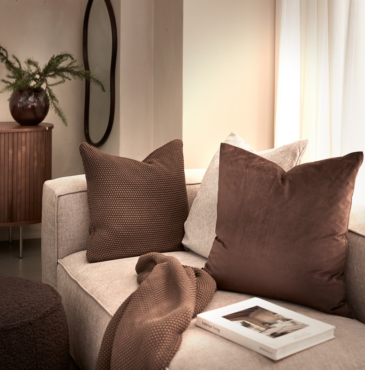 Beige sofa med brune puder, strikket plaid og en bog; træskab, bølget spejl og vase med grønt ved et lyst gardin.