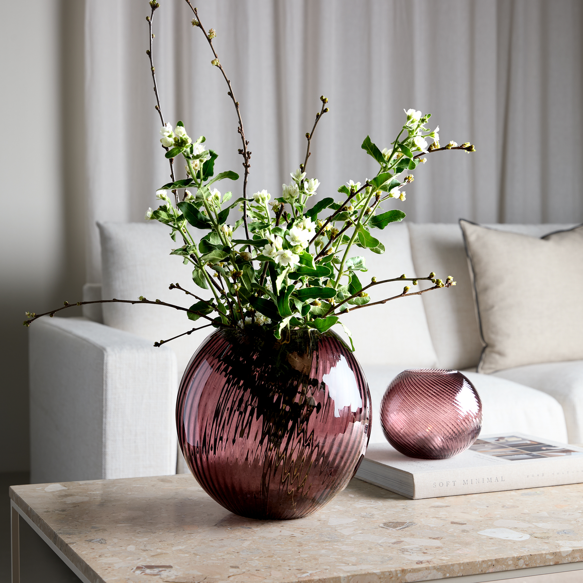 Rund rillet glasvase i mørk rosaton med grønne grene og hvide blomster på en stenlignende sofabordsplade; mindre matchende vase og en bog med titlen Soft Minimal ved lys, neutral stueindretning.