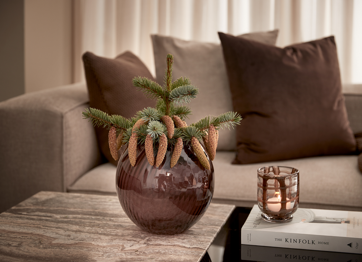 Rund brun glasvase med grene af gran og kogler på et marmorbord foran en beige sofa; en tænd fyrfadsstage står på to bøger, hvoraf den ene har titlen The Kinfolk Home.