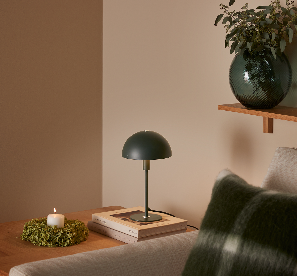 Grøn bordlampe på bøger ved siden af et tændt bloklys i grøn krans; sofa med grønternet pude og glasvase med grene på træhylde.
