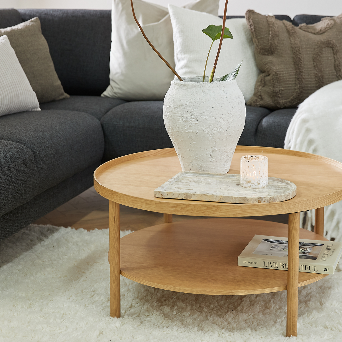 Rundt sofabord i lyst træ med en marmoragtig bakke, en hvid struktureret vase med grønne blade og en tændt lyslygte; på nederste hylde ligger en bog med titlen “LIVE BEAUTIFUL”, grå sofa med puder og blød hvidt tæppe i en lys stue.
